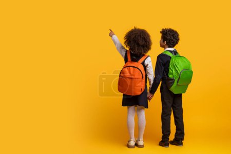 Téléchargez les photos : Des frères et sœurs afro-américains en tenue scolaire tenant la main sur un fond jaune dépeignent l'enfance et le lien entre les Noirs. espace de copie, vue arrière - en image libre de droit