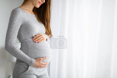 Una imagen suave que captura a una mujer embarazada con ropa casual, sosteniendo tiernamente su vientre, de pie junto a una ventana con cortinas blancas