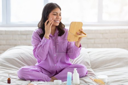 Téléchargez les photos : Femme du Moyen-Orient souriante en pyjama violet appliquant une lotion tout en tenant un miroir et assise sur un lit avec la lumière du jour - en image libre de droit