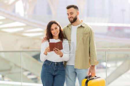 Foto de Una joven pareja sonriente de pie en un aeropuerto, con un pasaporte y una maleta amarilla, felices cónyuges milenarios listos para viajar de vacaciones, comprobando los documentos - Imagen libre de derechos