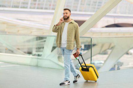 Photo for Smiling Man Talking On Phone While Walking With Suitcase At Airport Terminal, Happy Handsome Male Enjoying Pleasant Cellphone Conversation While Going To Departure Gate, Copy Space - Royalty Free Image
