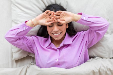 A muslim woman lying in bed touching her face and smiling, depicting a relatable morning scene at home