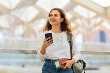 Cheerful young woman holding passport and smartphone while walking at airport, happy millennial female travelling alone, excited for upcoming trip, copy space