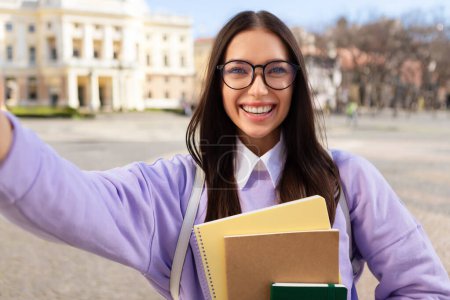 Téléchargez les photos : Une jeune étudiante heureuse portant des lunettes prend un selfie avec son téléphone, capturant le moment et la joie sur le campus de l'université - en image libre de droit