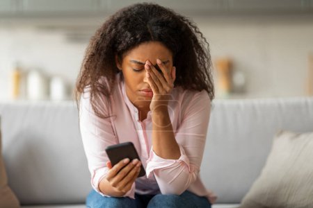Foto de Una señora negra aparece angustiada mientras mira la pantalla de su smartphone, sentada en casa con una mano en la frente - Imagen libre de derechos