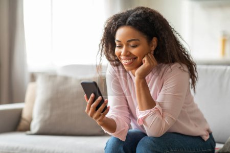 Téléchargez les photos : Une femme afro-américaine semble ravie alors qu'elle utilise son smartphone, assise confortablement sur le canapé à la maison - en image libre de droit