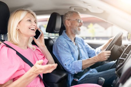 Photo for A retired married couple in their car, with the woman having a cheerful phone conversation as the senior man drives with a happy demeanor - Royalty Free Image