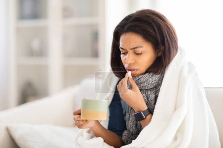 Eine kranke afrikanisch-amerikanische Dame ist in eine Decke gehüllt und scheint sich zu Hause krank zu fühlen und Tabletten zu nehmen