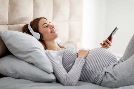Téléchargez les photos : Une future dame européenne se prélasse confortablement sur son lit, s'engage avec son téléphone, évoquant la chaleur de la maison et la sérénité de la grossesse - en image libre de droit