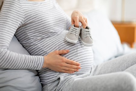 Foto de Mujer embarazada europea en casa, acunando su bulto y sosteniendo zapatos infantiles, representando los tiernos momentos de expectativa durante el embarazo - Imagen libre de derechos