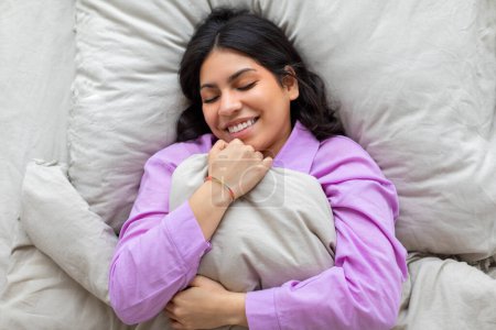 Content middle eastern woman in purple pajamas hugging a pillow with closed eyes and a smiling face, lying on a bed