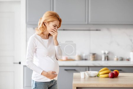 Foto de Una mujer embarazada europea está en su elegante cocina contemplando la nutrición y el cuidado prenatal, tiene náuseas matutinas - Imagen libre de derechos