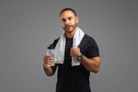Téléchargez les photos : Un homme concentré dans un t-shirt noir tient une bouteille d'eau et une serviette, indiquant une pause dans sa routine de remise en forme - en image libre de droit