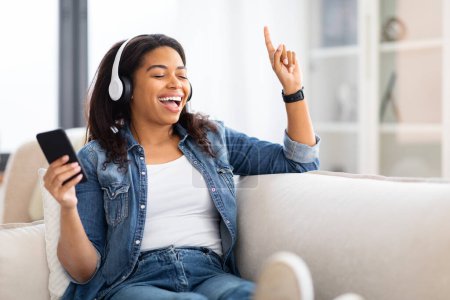 Téléchargez les photos : Une dame noire danse et chante avec joie tout en écoutant de la musique à la maison, en utilisant son smartphone et son casque - en image libre de droit