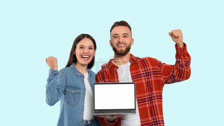 Téléchargez les photos : Couple excité affichant fièrement un écran d'ordinateur portable vierge, incarnant la jeunesse et le triomphe, sur un fond bleu - en image libre de droit