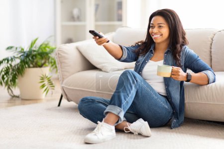 Foto de Una dama afroamericana relajada se sienta en casa sosteniendo un control remoto y una taza, probablemente disfrutando de un momento de ocio - Imagen libre de derechos