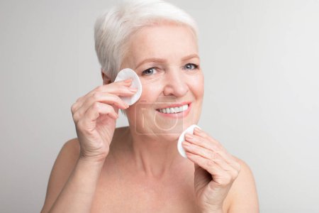 Une femme âgée européenne essuie son visage avec des tampons de coton, symbolisant les pratiques d'hygiène et d'autosoin au c?ur de s3niorlife