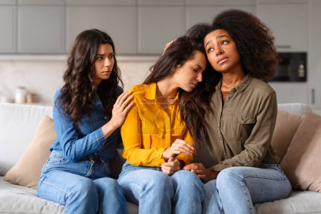 Téléchargez les photos : Confort et soutien à un ami, trois jeunes femmes multiraciales assises sur le canapé à la maison font preuve d'empathie - en image libre de droit