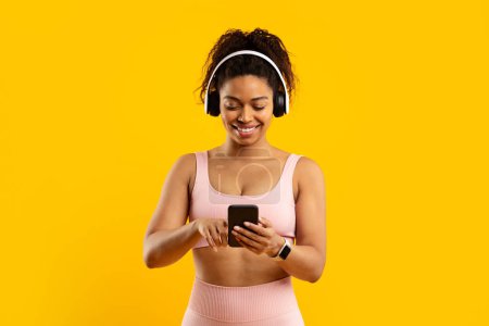 Téléchargez les photos : Joyeux dame noire sélectionnant la musique sur son téléphone tout en portant des écouteurs et des vêtements de fitness, isolé sur jaune - en image libre de droit