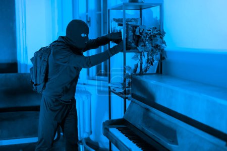 En un apartamento con poca luz, un ladrón es capturado robando objetos de valor de un gabinete, ilustrando un robo silencioso por la noche