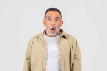 Téléchargez les photos : Un homme d'âge moyen avec une barbe grise montre une expression surprise, les yeux écarquillés et la bouche ouverte sur un fond gris - en image libre de droit