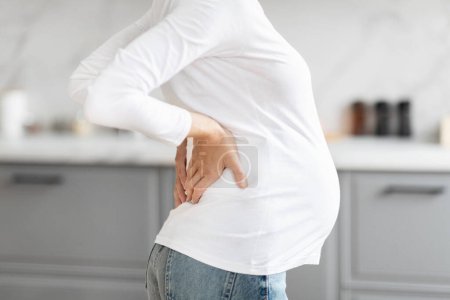 Foto de Mujer embarazada europea en una cocina blanca tiende a su espalda baja, consciente de su cuerpo cambiante, recortado - Imagen libre de derechos
