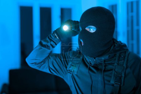 Foto de En plena noche, un ladrón vestido de negro escanea un apartamento antes de robar, su cara oscurecida por un pasamontañas en el contexto de una noche azul - Imagen libre de derechos