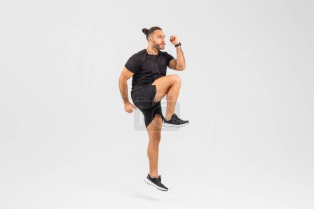 Téléchargez les photos : Un homme en forme en tenue de sport effectuant un exercice de jogging du genou haut à l'intérieur sur un fond blanc clair - en image libre de droit