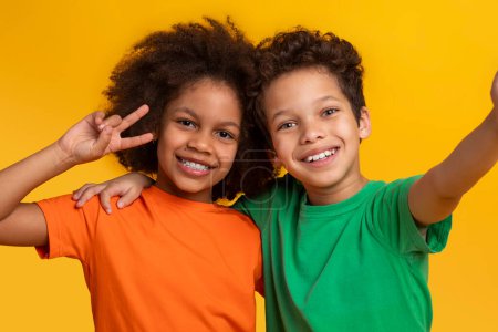 Téléchargez les photos : Frère et sœur afro-américains posent de façon ludique avec des gestes de paix dans un contexte jaune vif, reflétant l'esprit vif de l'enfance, prendre selfie - en image libre de droit