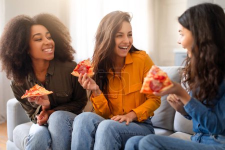 Drei junge multiethnische Freundinnen lachen und teilen Pizza in einer gemütlichen häuslichen Umgebung, die Wärme und Kameradschaft ausstrahlt