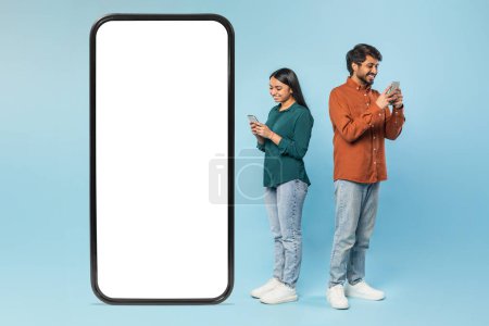 Téléchargez les photos : Homme et femme indiens penchés sur un écran géant de téléphone portable vierge adapté aux maquettes numériques sur bleu - en image libre de droit
