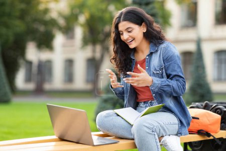 Photo for Exuding a sense of joy, an Indian lady laughs while looking at her laptop on a campus bench, highlighting the educational engagement and lively spirit of an eastern zoomer in an academic setting - Royalty Free Image
