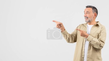 Photo for Cheerful old man pointing to the side, expressing positivity, wearing a beige shirt and t-shirt in an isolated studio setting, copy space - Royalty Free Image