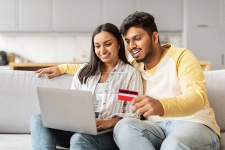 Foto de Esta imagen captura a una pareja india del milenio haciendo una compra en línea, con el marido sosteniendo una tarjeta de crédito, dentro de su ambiente acogedor hogar - Imagen libre de derechos