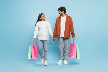 Indisches Paar mit Einkaufstaschen geht zusammen spazieren und tauscht warme, liebevolle Blicke auf blauem Hintergrund aus