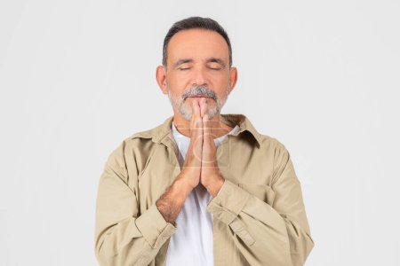 Foto de Anciano en un gesto de oración contemplativa, ojos cerrados, en camisa beige casual y camiseta blanca, aislado sobre un fondo blanco - Imagen libre de derechos