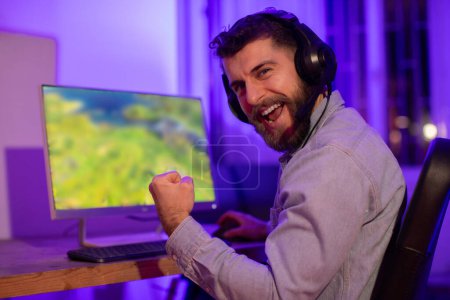 A cheerful man with headphones on raises his fist in victory while looking at a computer monitor, joy and excitement evident