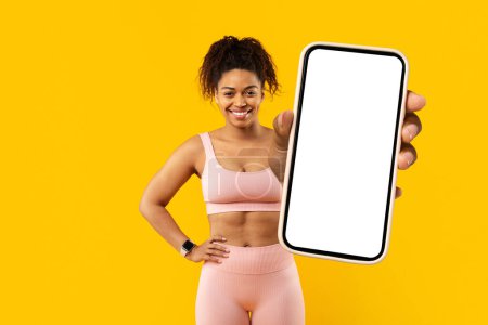 Téléchargez les photos : Une jeune femme afro-américaine en tenue de sport présente un écran de smartphone vierge sur un fond jaune vif, idéal pour le placement d'annonces - en image libre de droit