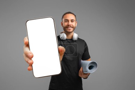 Photo for Man presenting a blank smartphone screen while holding a yoga mat, indicating an advertisement or app promo - Royalty Free Image