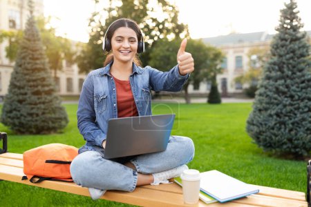 Photo for An Indian lady gives the thumbs up while using her laptop on a campus bench, symbolizing approval and positive feedback in a collegiate, setting as a tech-savvy zoomer with an educational focus - Royalty Free Image