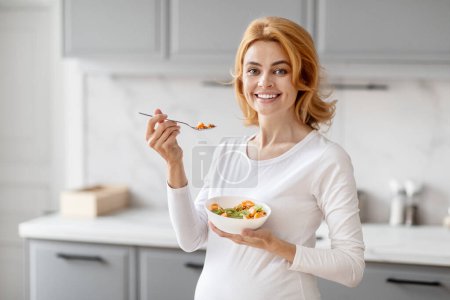Téléchargez les photos : Une femme enceinte européenne sourit dans une cuisine, tient un bol avec des aliments sains, promeut la nutrition - en image libre de droit