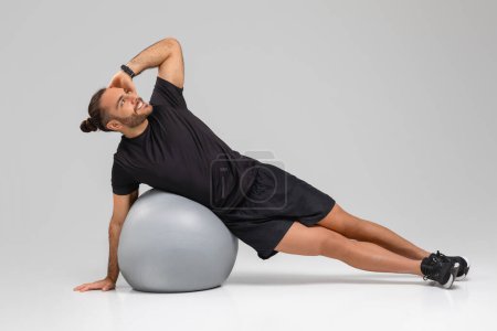 A focused guy in black sportswear maintaining balance on a grey exercise ball against a white backdrop