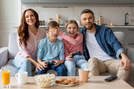 Love and playful mischief are evident as siblings cover each others eyes while the family enjoys video gaming at home, showcasing the dynamic of european family relationships