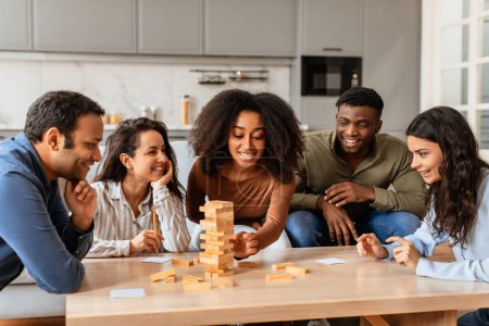 Photo for Multiracial young friends are seen focused and enjoying a block-stacking game together in a comfortable home setting - Royalty Free Image