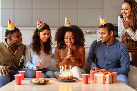 Eine geschlossene Gruppe junger multiethnischer Freunde versammelt sich um eine brennende Geburtstagstorte, bereit, im häuslichen Rahmen zu feiern