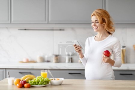 Foto de Una mujer europea expectante se absorbe en la lectura o la navegación en su teléfono inteligente, en una cocina bien iluminada con alimentos nutritivos a mano - Imagen libre de derechos