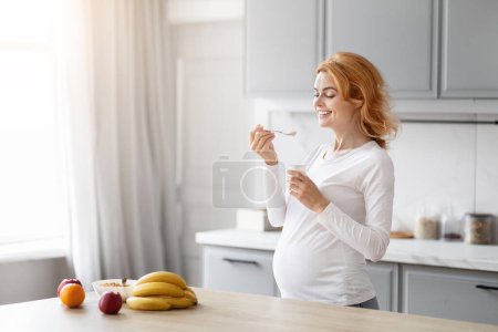 Foto de Mujer embarazada europea disfruta de un yogur nutritivo en su cocina moderna para apoyar su bienestar, espacio de copia - Imagen libre de derechos