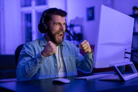 Excited bearded man cheers with clenched fists in front of computer screen, immersed in a neon-lit gaming session