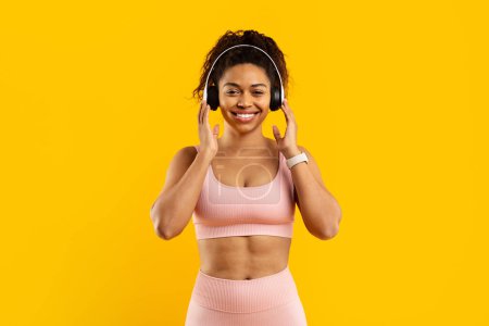 A smiling young african american woman wearing a pink sports bra and leggings with headphones against a vibrant yellow background enjoys music