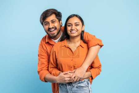 Ein lächelnder indischer Mann und eine Frau umarmen sich herzlich, beide tragen leuchtend orangefarbene Hemden vor blauem Hintergrund
